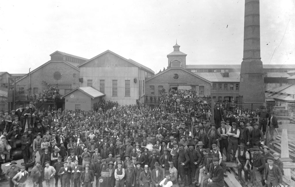 Black and white photograph of a crowd of men standing in front on a series of factory buildings. There is a large chimney on the right.